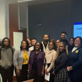 Group of diverse young professionals and students posing for a photo in front of a blue presentation screen in a conference space.