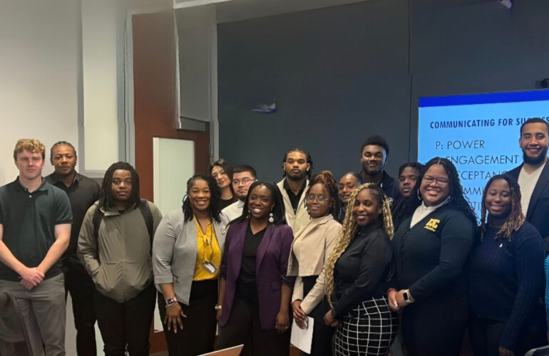 Group of diverse young professionals and students posing for a photo in front of a blue presentation screen in a conference space.