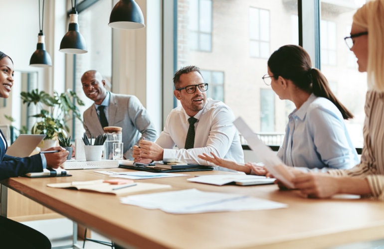 Diverse group of coworkers in a modern office discussing documents around a wooden conference table.